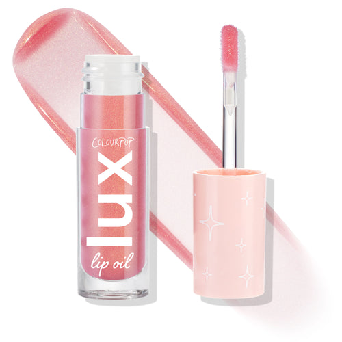 Comb By ColourPop Lux Lip Oil, bottle and open doe-foot applicator side by side, displaying a sheer shimmery pink swatch; clear tube labeled lux lip oil with a blush cap dotted with white starbursts, on a clean white background.