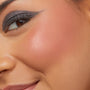 Cheek wearing ColourPop Cream Blush in shade Obviously, showing a soft peach flush with a dewy finish as the model smiles; smoky winged eye makeup visible in a tight close-up for product application detail.