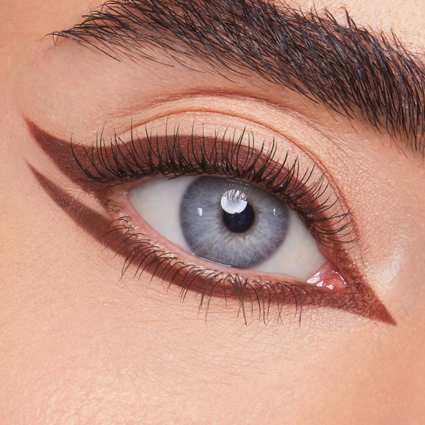 ColourPop Crème Gel Liner Sunnyvale tracing a sharp rich-brown double wing on a blue eye - lining upper and lower lashes with a pointed inner corner - over nude shadow, showcasing precise, long-wear crème gel eyeliner for graphic looks.