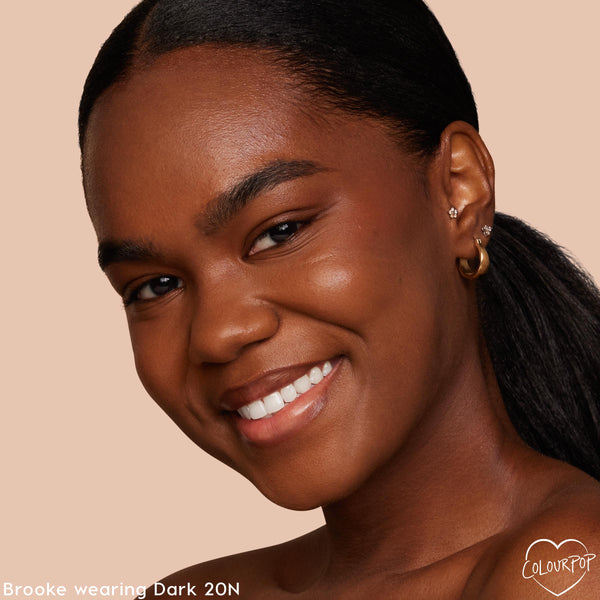 Model wearing ColourPop Pretty Fresh Tinted Moisturizer in Dark 20 N smiles in a close-up, showing dewy, even coverage and a natural glow; neutral beige backdrop, slicked-back hair, gold hoop and stud earrings.