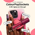 ColourPop Lip Stain tube snaps into a pink heart-shaped ColourPopSockets holder on a gradient pink smartphone, functioning as phone grip, stand, and lip holder while a hand with floral nail art grips the device - 3-in-1 on-the-go lip stain.