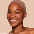 Model with close-cropped platinum hair smiles, showcasing ColourPop Pretty Fresh Creamy Concealer in shade Dark 158 W with smooth, radiant coverage; tight headshot on a soft beige background.