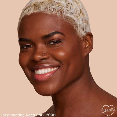 ColourPop Hyaluronic Hydrating Foundation in Deep Dark 200 N on a close-up smiling model, showing a smooth, dewy, even complexion with a neutral undertone against a warm beige studio background.