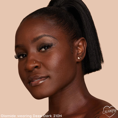 Model wearing ColourPop Pretty Fresh Creamy Concealer in Deep Dark 210 N, displaying smooth, even coverage and a neutral undertone on deep skin in a close-up portrait against a warm beige background.