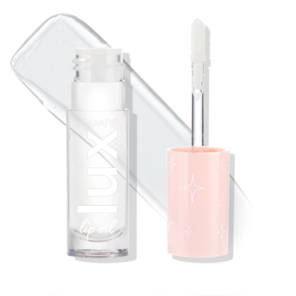 ColourPop Lux Lip Oil Dew Drop stands open, displaying a soft doe-foot applicator and transparent formula with a glossy swatch behind; clear square bottle and blush-pink cap with white star motifs on a bright white background.