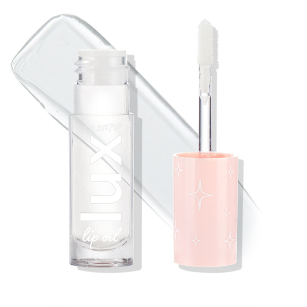 ColourPop Lux Lip Oil Dew Drop stands open, displaying a soft doe-foot applicator and transparent formula with a glossy swatch behind; clear square bottle and blush-pink cap with white star motifs on a bright white background.