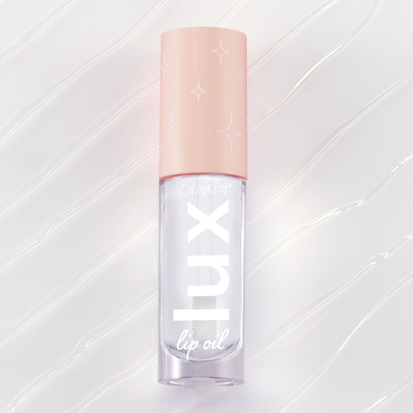 ColourPop Dew Drop Lux Lip Oil lying flat, a clear high-shine formula in a rectangular tube with blush-pink cap printed with starbursts and bold lux lettering, set on a pearlescent swirled backdrop for a glossy finish.