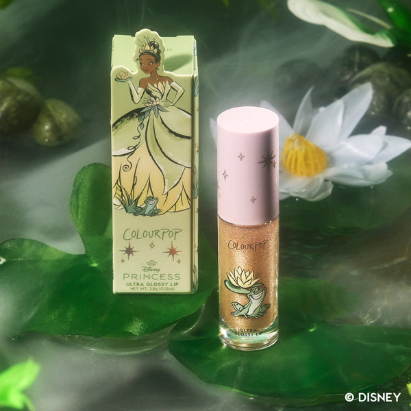 ColourPop Ambitious Tiana Ultra Glossy Lip from the Disney Princess collection standing next to its illustrated Tiana box, both glowing amid green lily pads and a white water lily in a dreamy, lagoon-inspired setting.