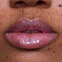 Close-up of freckled lips wearing Courageous Rapunzel Ultra Glossy Lip, a shimmering pink-lilac Disney Princess lip gloss with high-shine, iridescent sparkle on medium skin tone, showing smooth, hydrated finish and vivid color payoff.