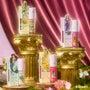 Princess Pout full collection set of Disney Princess ultra glossy lip glosses standing on ornate gold columns, featuring Ariel, Belle, Tiana, and Rapunzel packaging against a pink satin backdrop with flowers and moss, ColourPop collaboration.