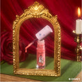 Mademoiselle Belle Ultra Glossy Lip from the Disney Princess collection stands open with a glittery coral applicator wand angled above the tube, framed by an ornate gold mirror beside a red rose and candle on a green surface, evoking Beauty and the Beast.
