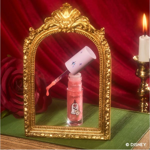 Mademoiselle Belle Ultra Glossy Lip from the Disney Princess collection stands open with a glittery coral applicator wand angled above the tube, framed by an ornate gold mirror beside a red rose and candle on a green surface, evoking Beauty and the Beast.