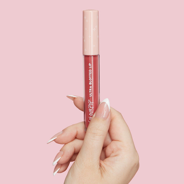 ColourPop Doozy Ultra Blotted Lip held upright by a manicured hand, showing a rosy pink stain in a clear tube with white logo and a blush cap dotted with stars, set against a pastel pink background for a clean product shot.