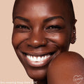 Smiling model with deep brown skin wears ColourPop Hyaluronic Hydrating Foundation in Deep Dark 213 C, showing smooth, dewy, even coverage; close-up face with nose ring and ear piercings against a warm beige background.