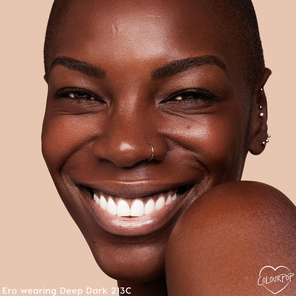 Smiling model with deep brown skin wears ColourPop Hyaluronic Hydrating Foundation in Deep Dark 213 C, showing smooth, dewy, even coverage; close-up face with nose ring and ear piercings against a warm beige background.