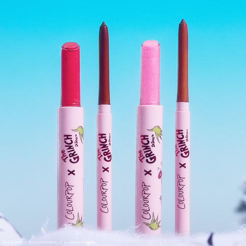 Colourpop Pouty Grinch lip set - four slim pink tubes with The Grinch x Colourpop branding - standing upright, showcasing a red lipstick, two neutral liners, and a shimmery pink balm on a snowy base against a bright blue sky.