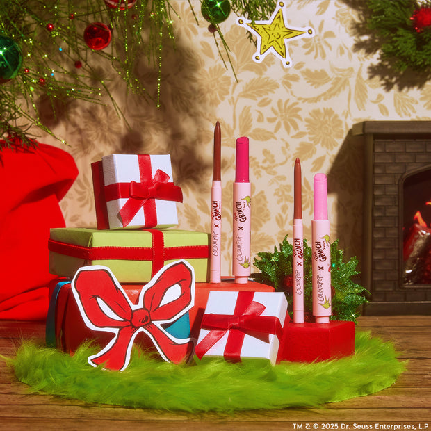 Pouty Grinch lip set by ColourPop stands upright on wrapped gifts - two Lippie Stix and two Lippie Pencils in pink and nude shades - styled with red bows, green faux fur, and a cozy fireplace for a festive holiday scene.