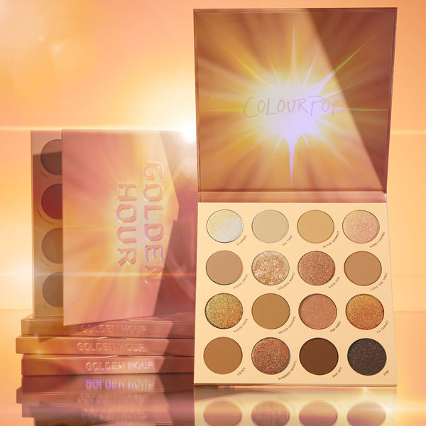 Golden Hour ColourPop Shadow Palette displaying a 16-pan mix of warm mattes, metallics, and glitter, lid with sunburst art propped open, with stacked palettes beside it on a reflective surface in a glowing golden studio.
