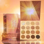 Golden Hour ColourPop Shadow Palette displaying a 16-pan mix of warm mattes, metallics, and glitter, lid with sunburst art propped open, with stacked palettes beside it on a reflective surface in a glowing golden studio.