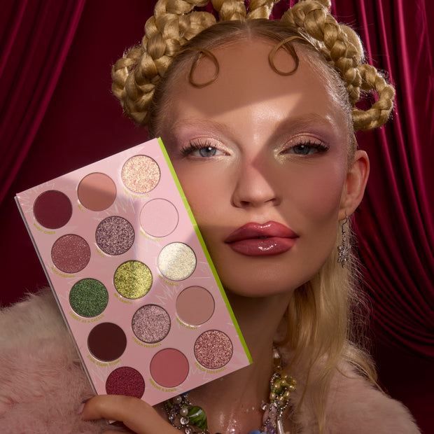 How the Grinch Stole Christmas Shadow Palette by ColourPop held beside a person’s glam face, showing a pink 16-pan mix of matte and glitter eyeshadows - greens, pinks, golds, and plums - set against rich red drapery for a festive holiday look.