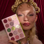 How the Grinch Stole Christmas Shadow Palette by ColourPop held beside a person’s glam face, showing a pink 16-pan mix of matte and glitter eyeshadows - greens, pinks, golds, and plums - set against rich red drapery for a festive holiday look.