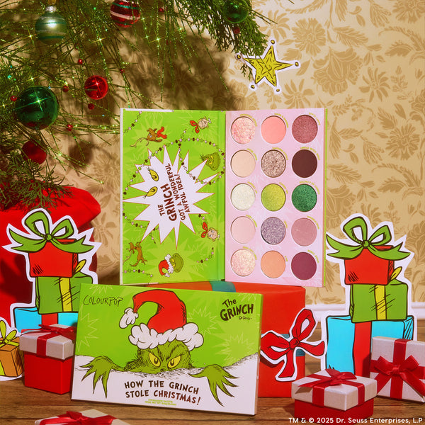 ColourPop x The Grinch eyeshadow palette opens to 15 holiday shades - pinks, greens, and golds in matte and shimmer - displayed with Grinch artwork, set among Christmas ornaments and wrapped gifts for a festive makeup scene.