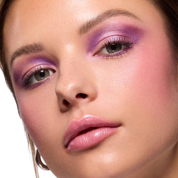 Violet and lilac shimmer blended across lids on a model, showcasing the ColourPop It’s My Pleasure shadow palette; close-up beauty shot with soft pink blush, defined lashes, and glossy pink lips on dewy skin.