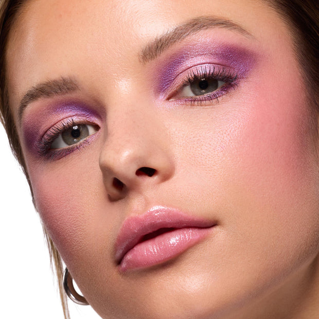 Violet and lilac shimmer blended across lids on a model, showcasing the ColourPop It’s My Pleasure shadow palette; close-up beauty shot with soft pink blush, defined lashes, and glossy pink lips on dewy skin.