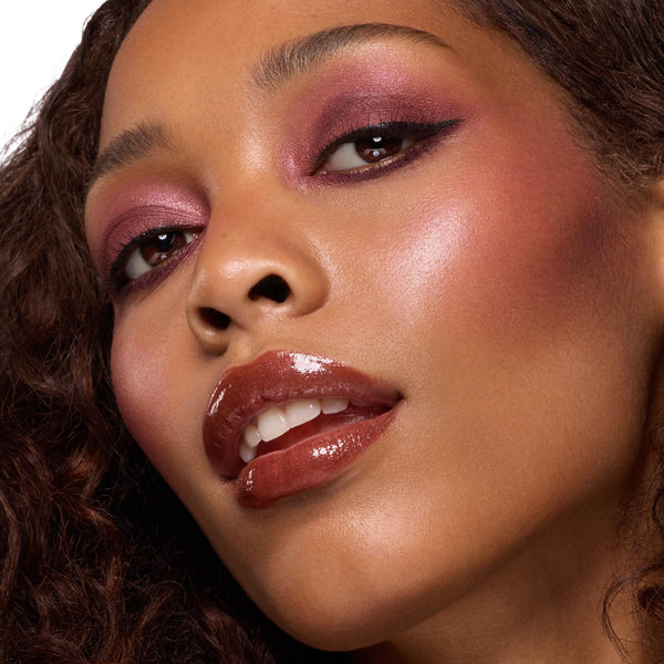 Close-up of high-shine lips wearing ColourPop So Juicy Plumping Gloss Balm with Peptides in shade L-O-V-E, creating a fuller, glassy finish; model tilts head with warm brown gloss and wine-toned eyeshadow under studio lighting.