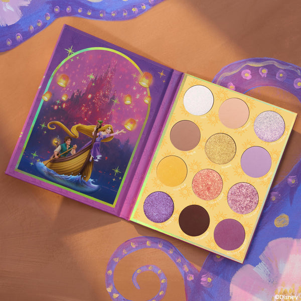 Best Day Ever ColourPop Shadow Palette lies open, showcasing a 12-pan mix of matte and shimmer lilac, gold, pink, taupe, and deep brown shades, with Disney Tangled Rapunzel lantern scene artwork on the left against a painted purple-and-gold background.