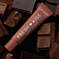ColourPop Fresh Kiss glossy lip balm tube from the Get Rich ColourPop Glossy Lip Balm Trio lying diagonally on milk and dark chocolate bars, terracotta-brown packaging labeled Fresh Kiss Glossy Lip Balm and ColourPop in a chocolate-themed flat lay.