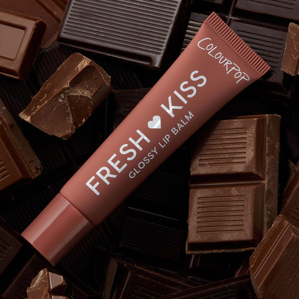 ColourPop Fresh Kiss glossy lip balm tube from the Get Rich ColourPop Glossy Lip Balm Trio lying diagonally on milk and dark chocolate bars, terracotta-brown packaging labeled Fresh Kiss Glossy Lip Balm and ColourPop in a chocolate-themed flat lay.
