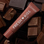 ColourPop Fresh Kiss glossy lip balm tube from the Get Rich ColourPop Glossy Lip Balm Trio lying diagonally on milk and dark chocolate bars, terracotta-brown packaging labeled Fresh Kiss Glossy Lip Balm and ColourPop in a chocolate-themed flat lay.