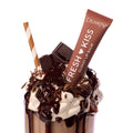 ColourPop Fresh Kiss Glossy Balm, shade Chocolate Milkshake, propped in a whipped-cream–topped chocolate milkshake with hot fudge, chocolate shavings, and a striped straw on a white studio background.