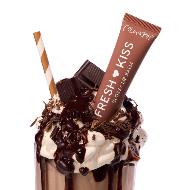 ColourPop Fresh Kiss Glossy Balm, shade Chocolate Milkshake, propped in a whipped-cream–topped chocolate milkshake with hot fudge, chocolate shavings, and a striped straw on a white studio background.
