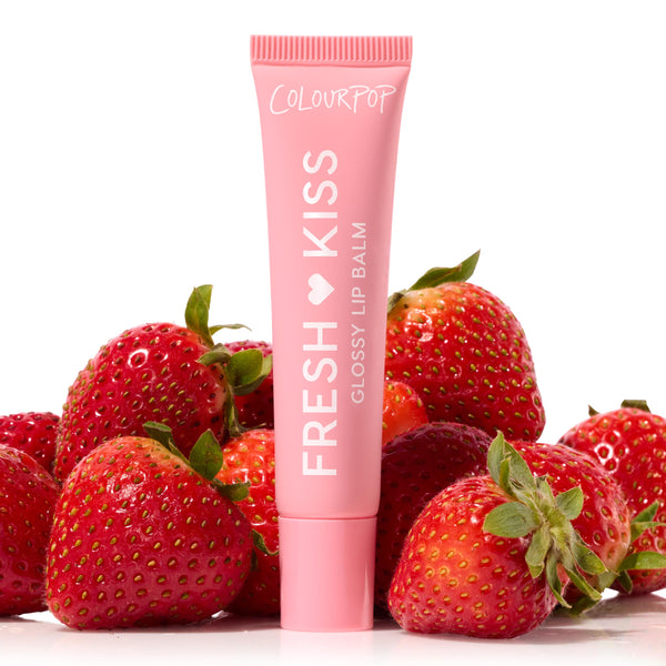 ColourPop Fresh Kiss Glossy Balm, shade Strawberry Milk, stands upright among fresh strawberries on a white backdrop, the pink squeeze tube labeled FRESH KISS Glossy Lip Balm, showcasing the fruit-inspired color and hydrating glossy finish.