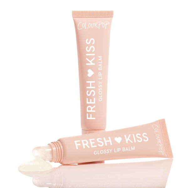 Two soft-pink tubes of ColourPop Fresh Kiss Glossy Balm in Vanilla Cake are arranged on a reflective white surface, one showing a bead of clear, glossy balm pooling from its slanted applicator tip.