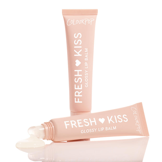 Two soft-pink tubes of ColourPop Fresh Kiss Glossy Balm in Vanilla Cake are arranged on a reflective white surface, one showing a bead of clear, glossy balm pooling from its slanted applicator tip.