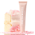 ColourPop Fresh Kiss Glossy Balm, Vanilla Cake, stands upright against a frosted vanilla cake slice topped with pink sprinkles, blush tube with white lettering styled on a bright, reflective surface for a sweet, high-shine lip finish.
