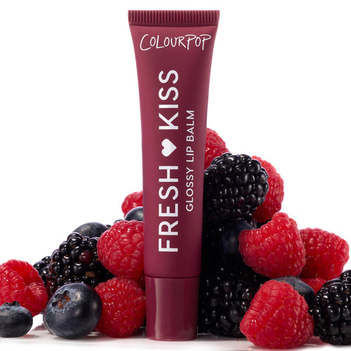 ColourPop Fresh Kiss Glossy Balm in Wild Berry standing upright, deep berry tube with white lettering nestled among fresh raspberries, blackberries, and blueberries on a white backdrop, highlighting juicy, glossy lip care.
