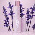 A-List Babe Serum Stick standing uncapped with dewy droplets on a pastel lavender tube, framed by purple lavender sprigs on a glossy pink surface; ColourPop Serum Stick by Fourth Ray Beauty for easy swipe-on face serum.