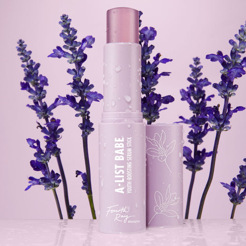 A-List Babe Serum Stick standing uncapped with dewy droplets on a pastel lavender tube, framed by purple lavender sprigs on a glossy pink surface; ColourPop Serum Stick by Fourth Ray Beauty for easy swipe-on face serum.