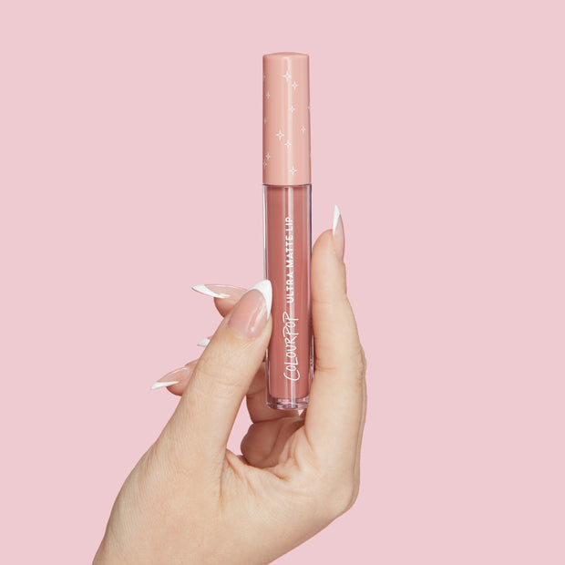 ColourPop Ultra Matte Lip Fifth Avenue tube held upright by a hand with a French manicure, showing a neutral nude-pink shade in a clear vial with a blush cap and white stars, set against a solid pastel pink background.