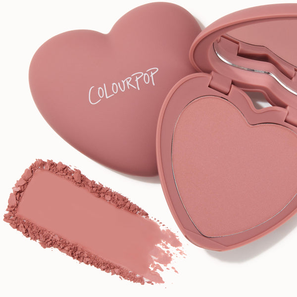 Heart-shaped compact displays ColourPop Pressed Powder Blush in Flirt Alert, a dusty rose pink, with the lid logo visible, an open mirror, and a crushed swatch smeared beside the case on a white background.