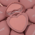 Heart-shaped Flirt Alert compact opens to reveal a soft peach‑pink pan and mirror, highlighting ColourPop Pressed Powder Blush; surrounded by matching dusty‑rose cases with ColourPop logos in a clean studio flat lay.