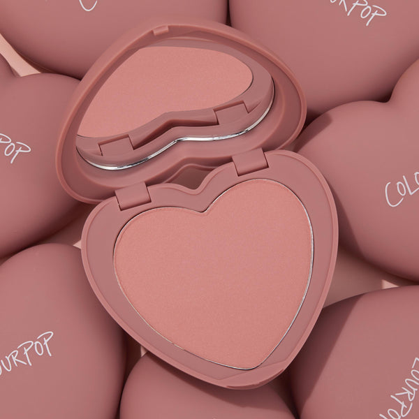 Heart-shaped Flirt Alert compact opens to reveal a soft peach‑pink pan and mirror, highlighting ColourPop Pressed Powder Blush; surrounded by matching dusty‑rose cases with ColourPop logos in a clean studio flat lay.