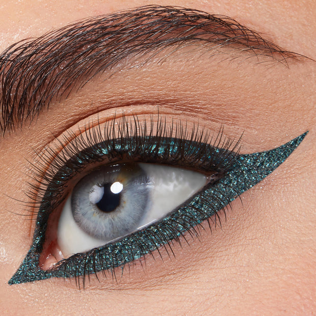 ColourPop Graphix Ink Liner in Forever defining a blue eye with a bold, elongated cat-eye; metallic teal shimmer lines upper and lower lash lines in a razor-sharp wing, framed by natural lashes and a groomed brow in a close-up.