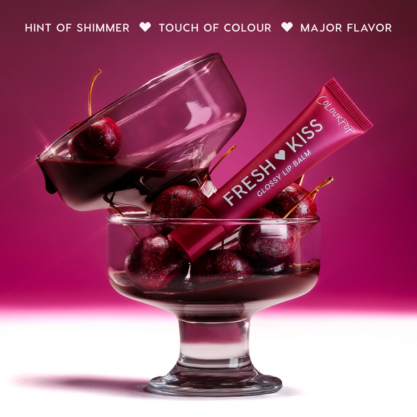 Colourpop Fresh Kiss Glossy Balm Chocolate Cherry tube squeezed open over a glass dessert dish filled with glossy cherries, showcasing rich berry shade and shimmering texture for Lip Gloss & Lip Oil lovers against a deep red background.