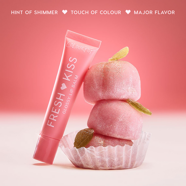Colourpop Fresh Kiss Glossy Balm in Peach Mochi leaning against stacked pink mochi candies with sugar crystals, highlighting the shimmery Lip Gloss & Lip Oil hybrid on a soft red and white background.