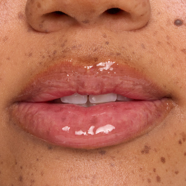 Glossy, hydrated lips wear a clear lip mask with small white balm flecks melting on the surface, shown in an extreme close-up on freckled skin to showcase ColourPop lip mask treatment shine and texture.
